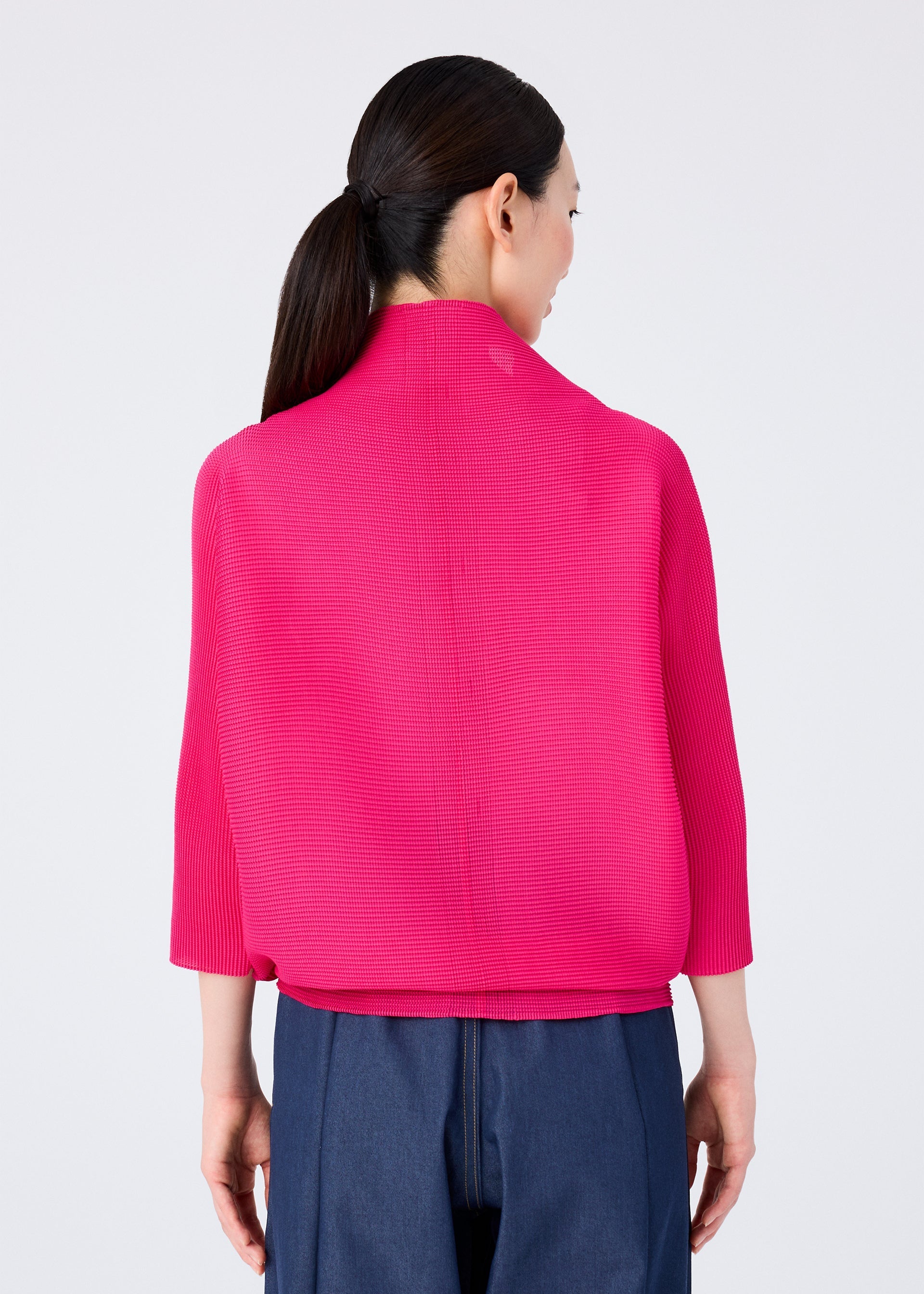 Zoomed view A woman with long dark hair in a low ponytail wears the me ISSEY MIYAKE DOLMAN STRETCH PLEATS 1, a bright pink 3/4 sleeve pleated cardigan, and dark blue high-waisted pants. She faces away against a plain light background.