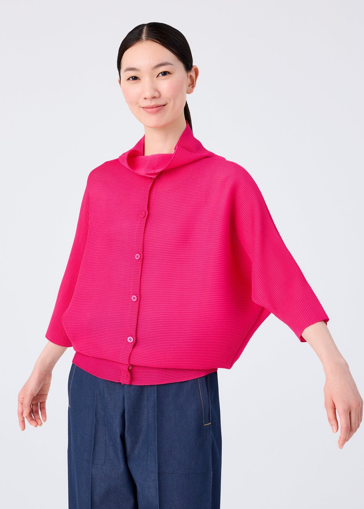 A woman with dark hair pulled back is smiling, wearing the me ISSEY MIYAKE DOLMAN STRETCH PLEATS 1 bright pink cardigan with wide sleeves and blue pants, standing against a plain white background. view 7 of 8
