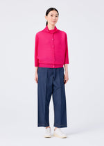 A woman stands against a white background wearing the me ISSEY MIYAKE DOLMAN STRETCH PLEATS 1 bright pink cardigan, loose blue wide-leg pants, and white slip-on shoes. She looks forward with a soft smile.