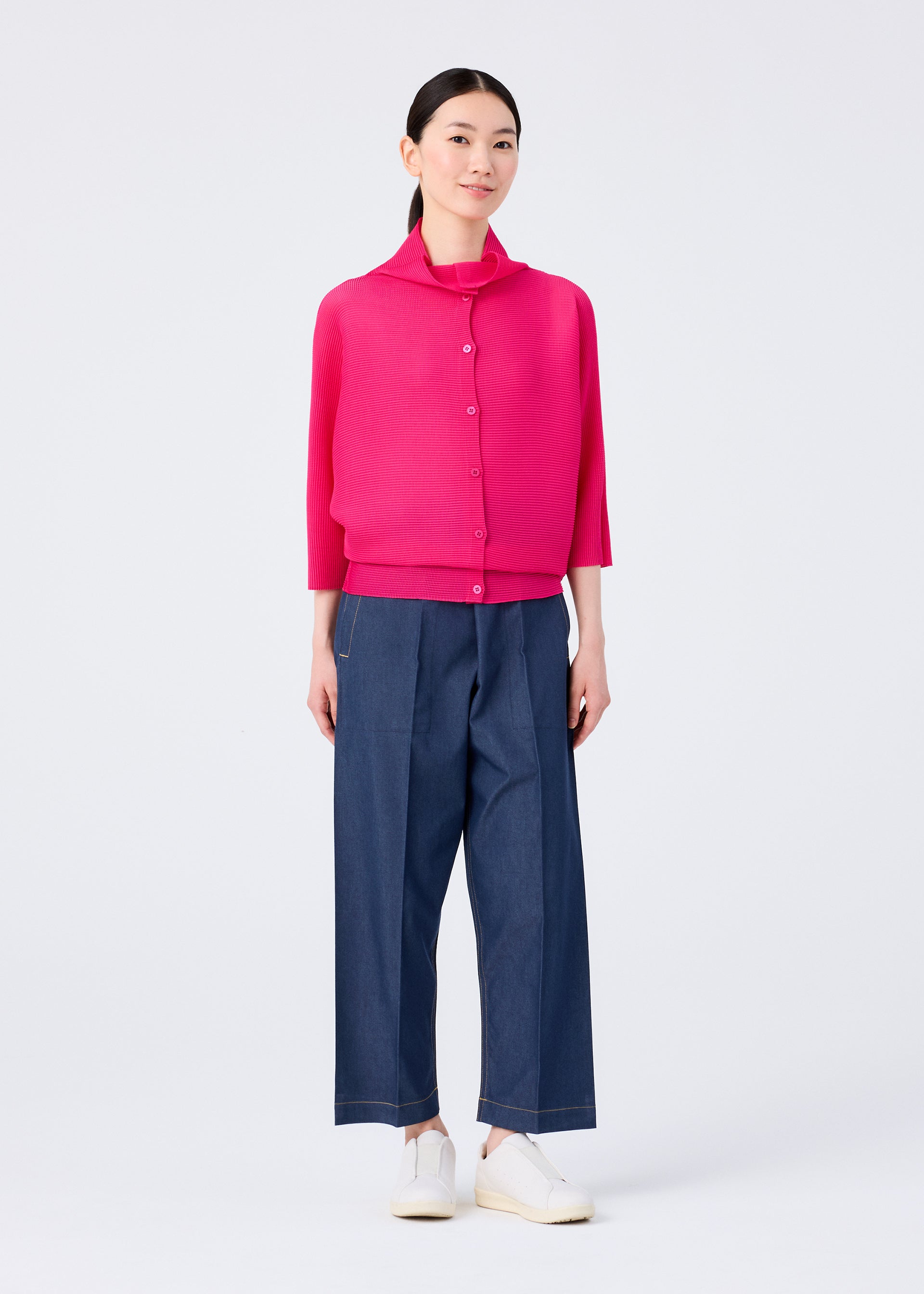 Zoomed view A woman stands against a white background wearing the me ISSEY MIYAKE DOLMAN STRETCH PLEATS 1 bright pink cardigan, loose blue wide-leg pants, and white slip-on shoes. She looks forward with a soft smile.