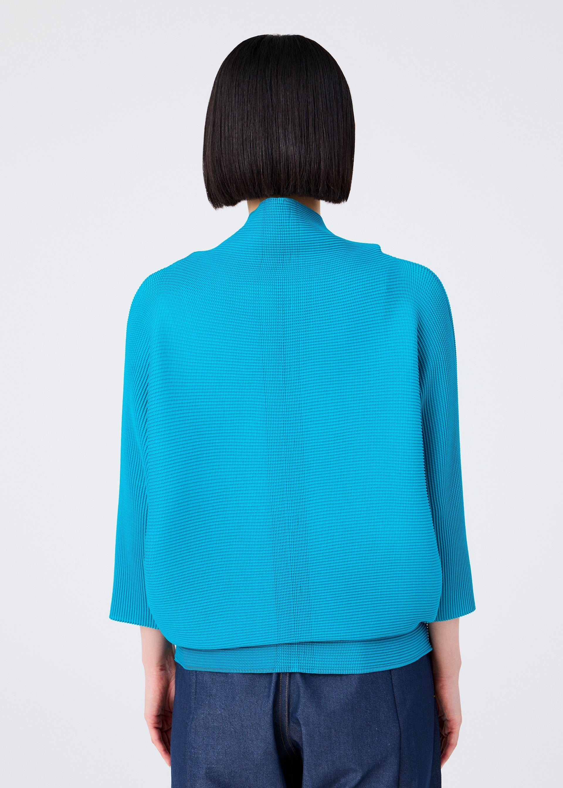 Zoomed view Seen from behind against a plain light background, a person with a straight bob wears the me ISSEY MIYAKE DOLMAN STRETCH PLEATS 2 cardigan in bright turquoise with three-quarter sleeves, paired with dark blue pants.