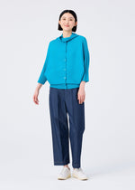 A woman smiles against a white background, wearing the me ISSEY MIYAKE DOLMAN STRETCH PLEATS 2 top in turquoise, paired with dark blue wide-leg pants and white shoes.