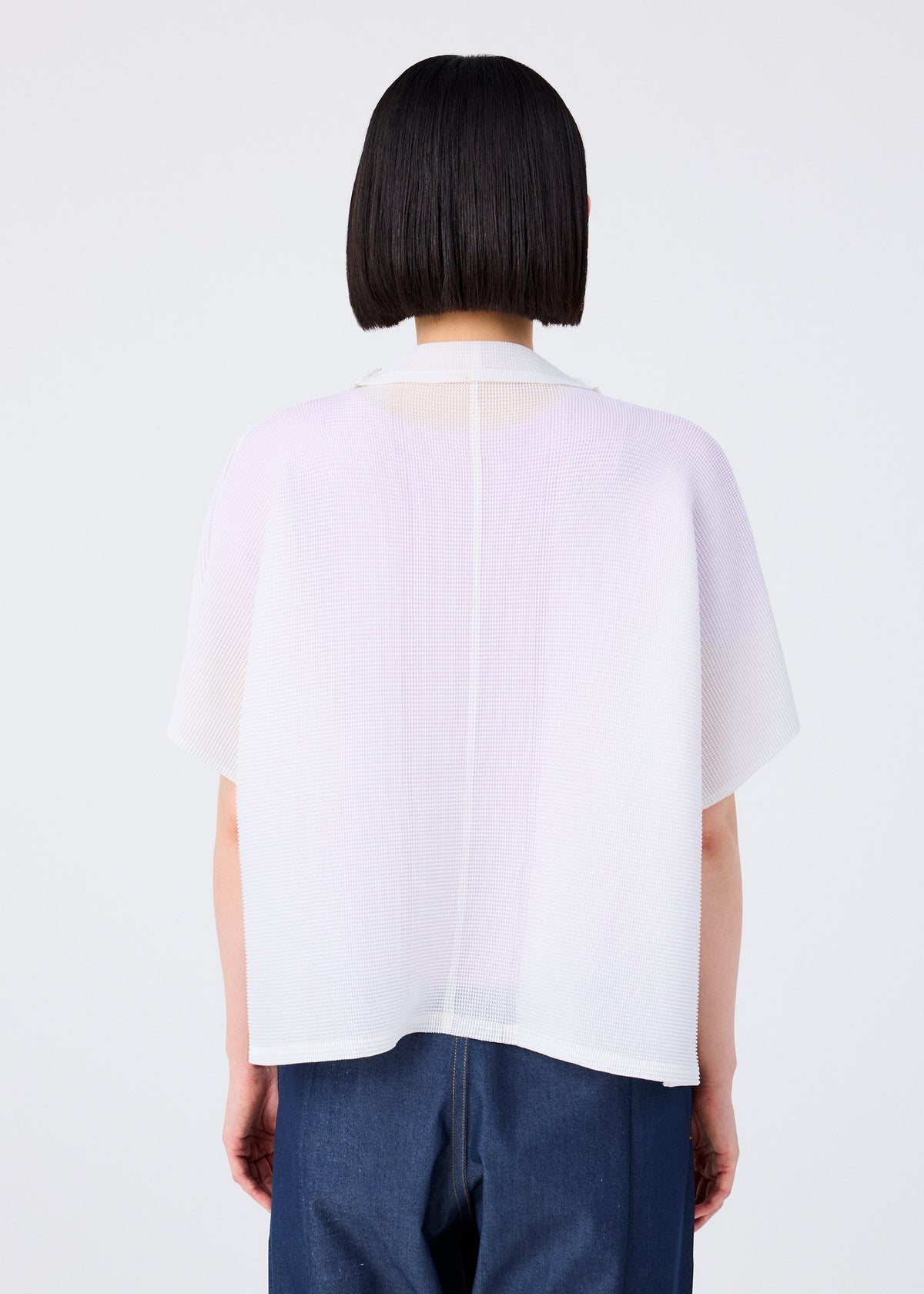 A person with straight, chin-length dark hair is seen from behind in a sheer white SQUARE STRETCH PLEATS top by me ISSEY MIYAKE over blue pants, creating a soft, three-dimensional silhouette against a plain light background. view 6 of 7