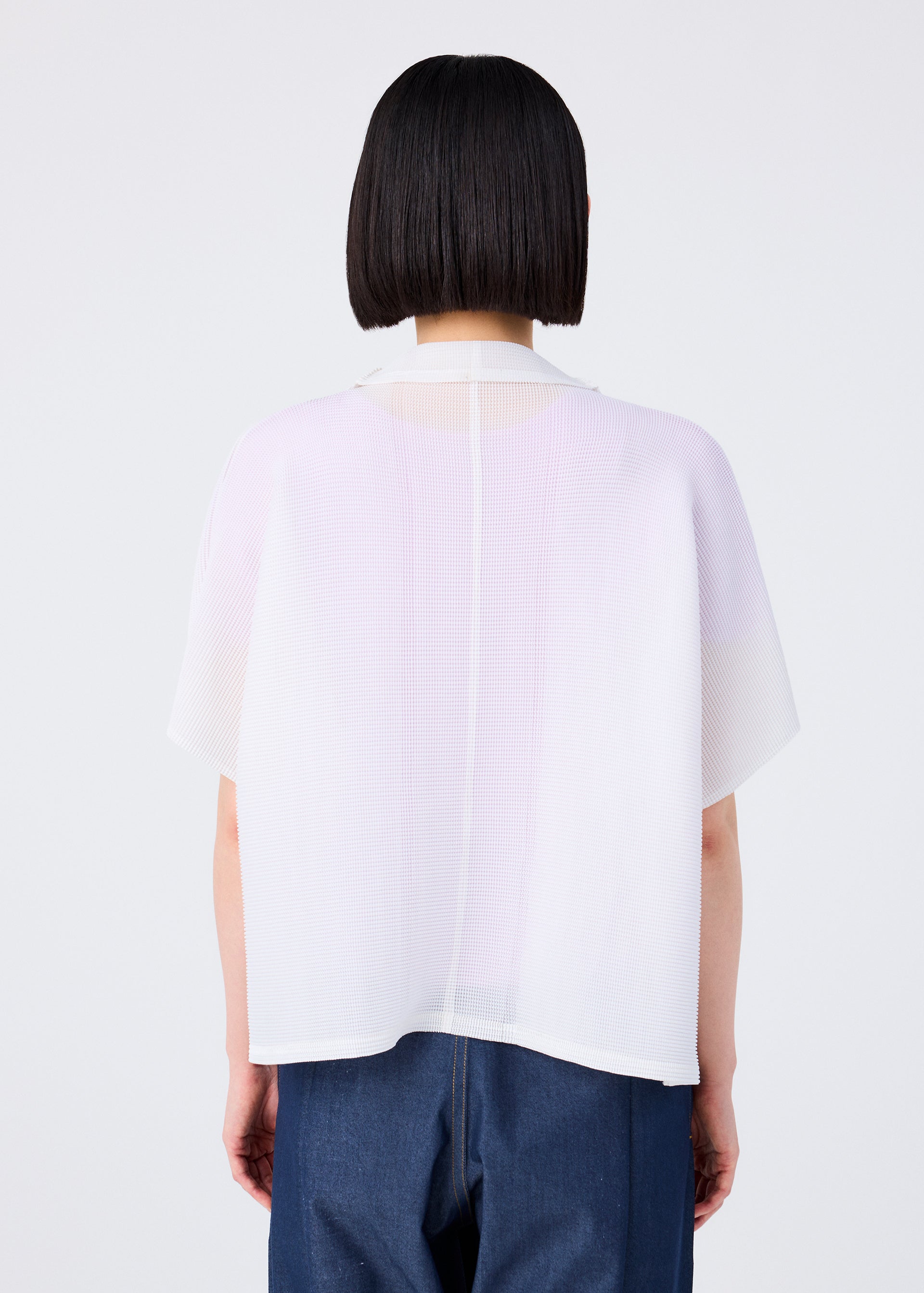 Zoomed view A person with straight, chin-length dark hair is seen from behind in a sheer white SQUARE STRETCH PLEATS top by me ISSEY MIYAKE over blue pants, creating a soft, three-dimensional silhouette against a plain light background.