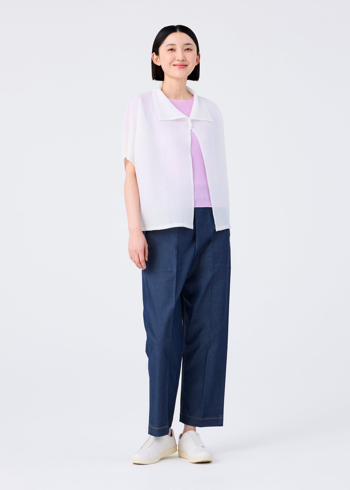 A person with straight dark hair wears a light purple top, a white short-sleeved cardigan, me ISSEY MIYAKE’s SQUARE STRETCH PLEATS blue wide-leg pants, and white slip-on shoes, standing against a plain white background and smiling slightly. view 7 of 7