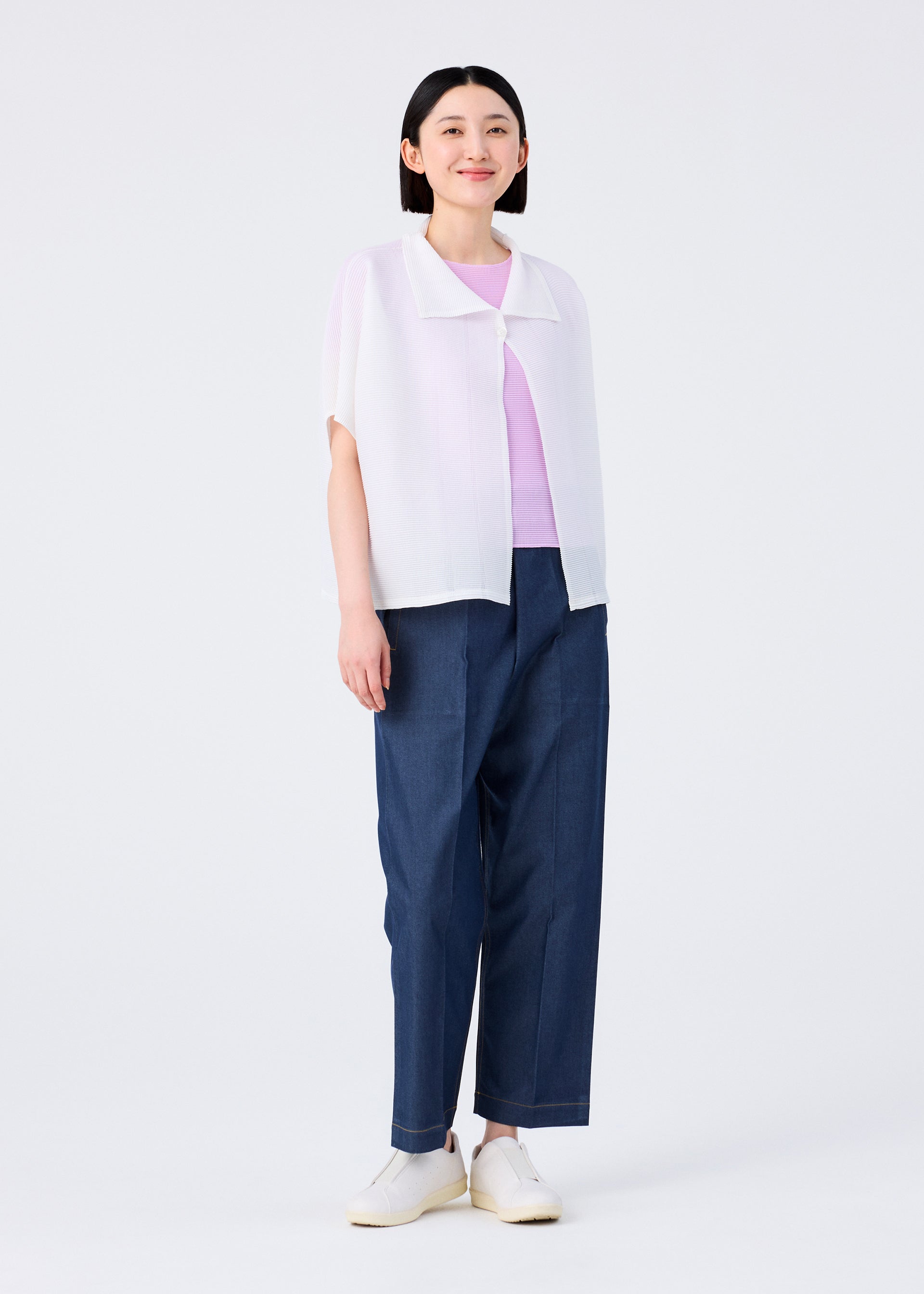 Zoomed view A person with straight dark hair wears a light purple top, a white short-sleeved cardigan, me ISSEY MIYAKE’s SQUARE STRETCH PLEATS blue wide-leg pants, and white slip-on shoes, standing against a plain white background and smiling slightly.