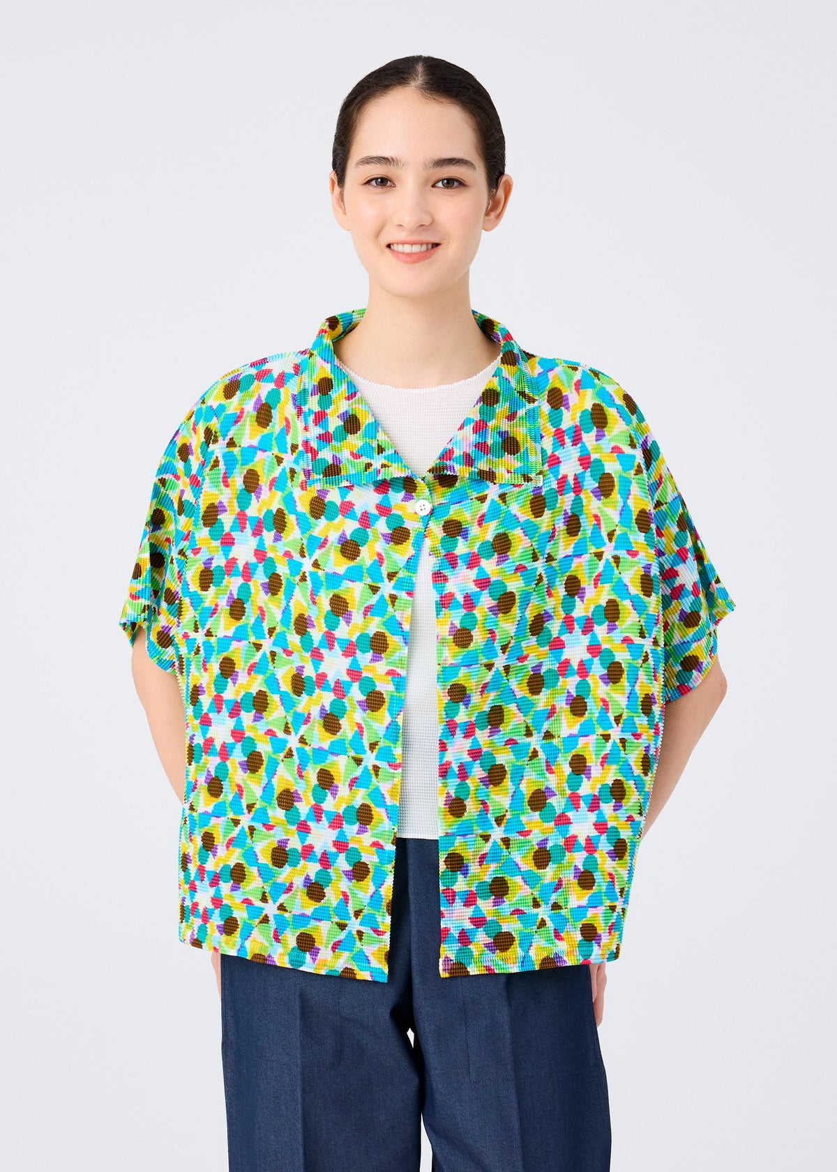 A woman smiles facing forward, wearing the me ISSEY MIYAKE KALEIDOSCOPE short-sleeved cardigan with a vibrant pattern over a white shirt and blue pants, set against a plain light background. view 4 of 6