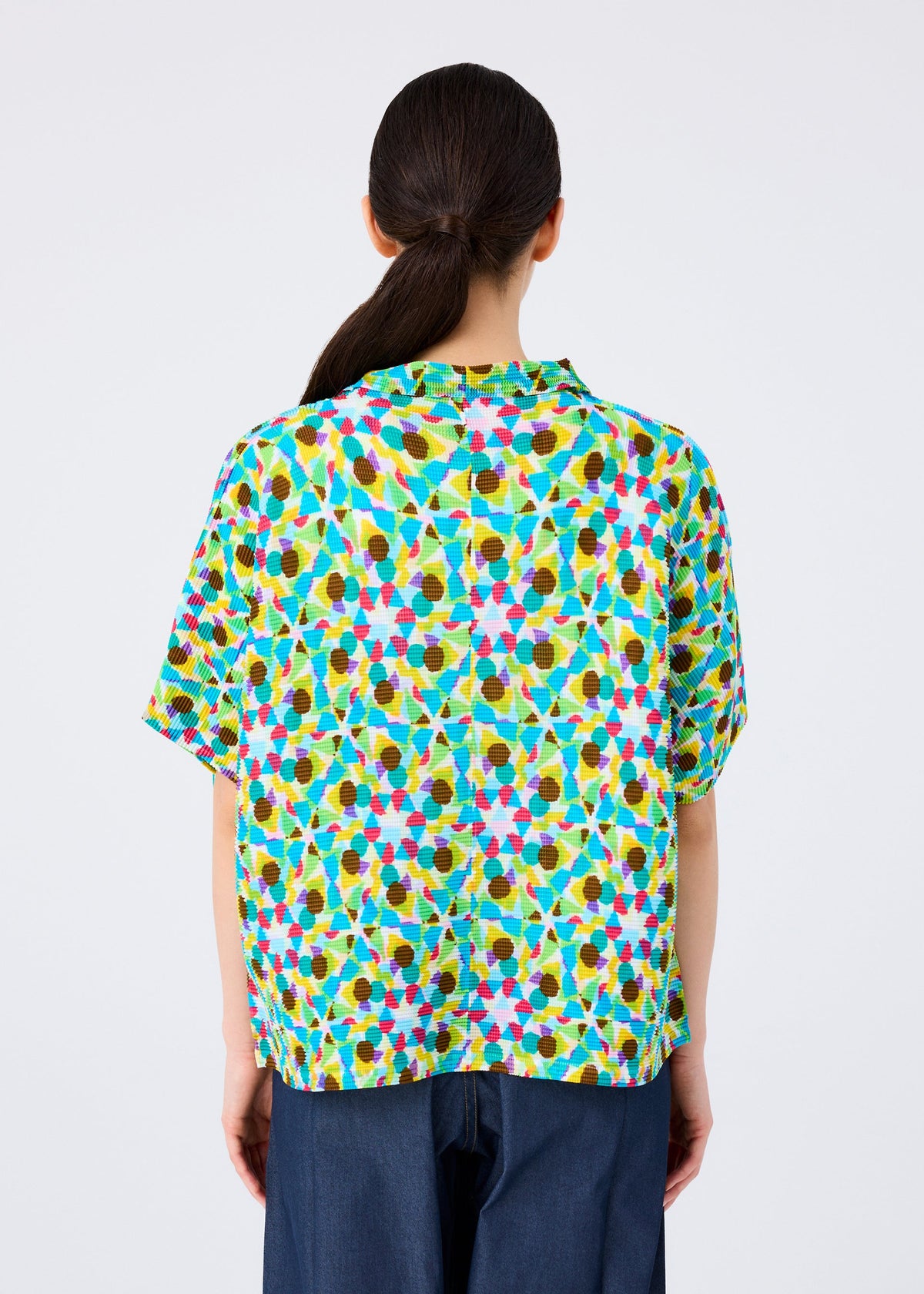 A person with long dark hair in a low ponytail is seen from behind, wearing the KALEIDOSCOPE short-sleeved cardigan by me ISSEY MIYAKE in green, yellow, pink, blue, and brown, paired with dark blue pants. view 5 of 6