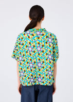 A person with long dark hair in a low ponytail is seen from behind, wearing the KALEIDOSCOPE short-sleeved cardigan by me ISSEY MIYAKE in green, yellow, pink, blue, and brown, paired with dark blue pants.