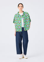 A person stands against a plain white background wearing the KALEIDOSCOPE short-sleeved cardigan by me ISSEY MIYAKE over a white top, paired with loose blue pants and white shoes.
