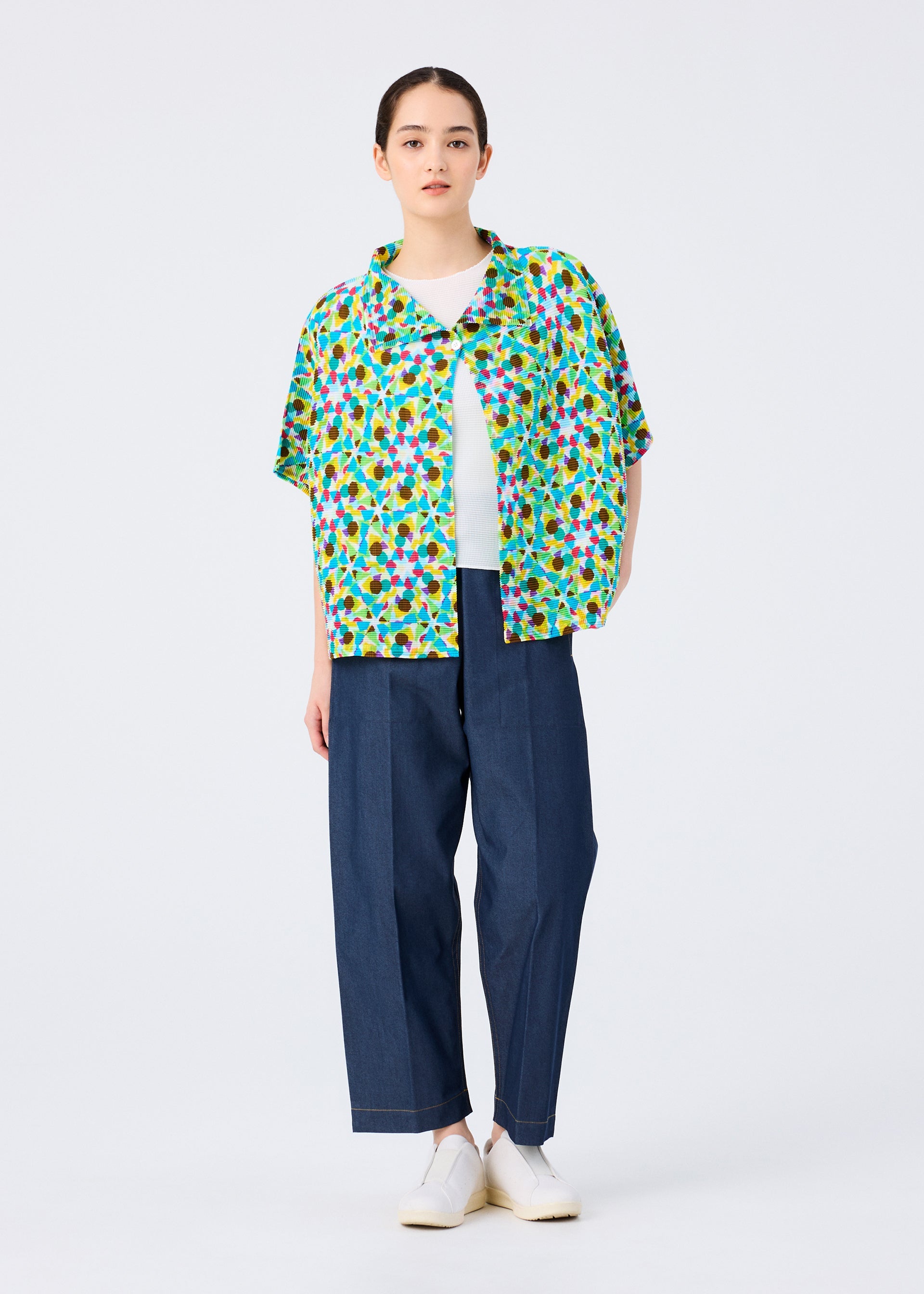 Zoomed view A person stands against a plain white background wearing the KALEIDOSCOPE short-sleeved cardigan by me ISSEY MIYAKE over a white top, paired with loose blue pants and white shoes.
