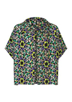 The KALEIDOSCOPE short-sleeved shirt by me ISSEY MIYAKE features a bold abstract floral print in green, yellow, purple, black, and beige on white. Shown laid flat with the back facing up. - BLACK-HUED （no.18）