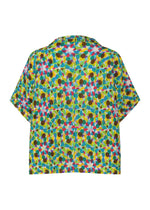 The KALEIDOSCOPE short-sleeve shirt by me ISSEY MIYAKE features a vibrant pattern of overlapping circles and hexagons in blue, green, pink, yellow, and brown. The shirt is laid flat to display the back design.