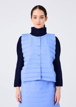 A woman with dark hair tied back models the RIPPLE PLEATS light blue padded vest and matching skirt by me ISSEY MIYAKE over a dark long-sleeve top, standing against a plain light background.