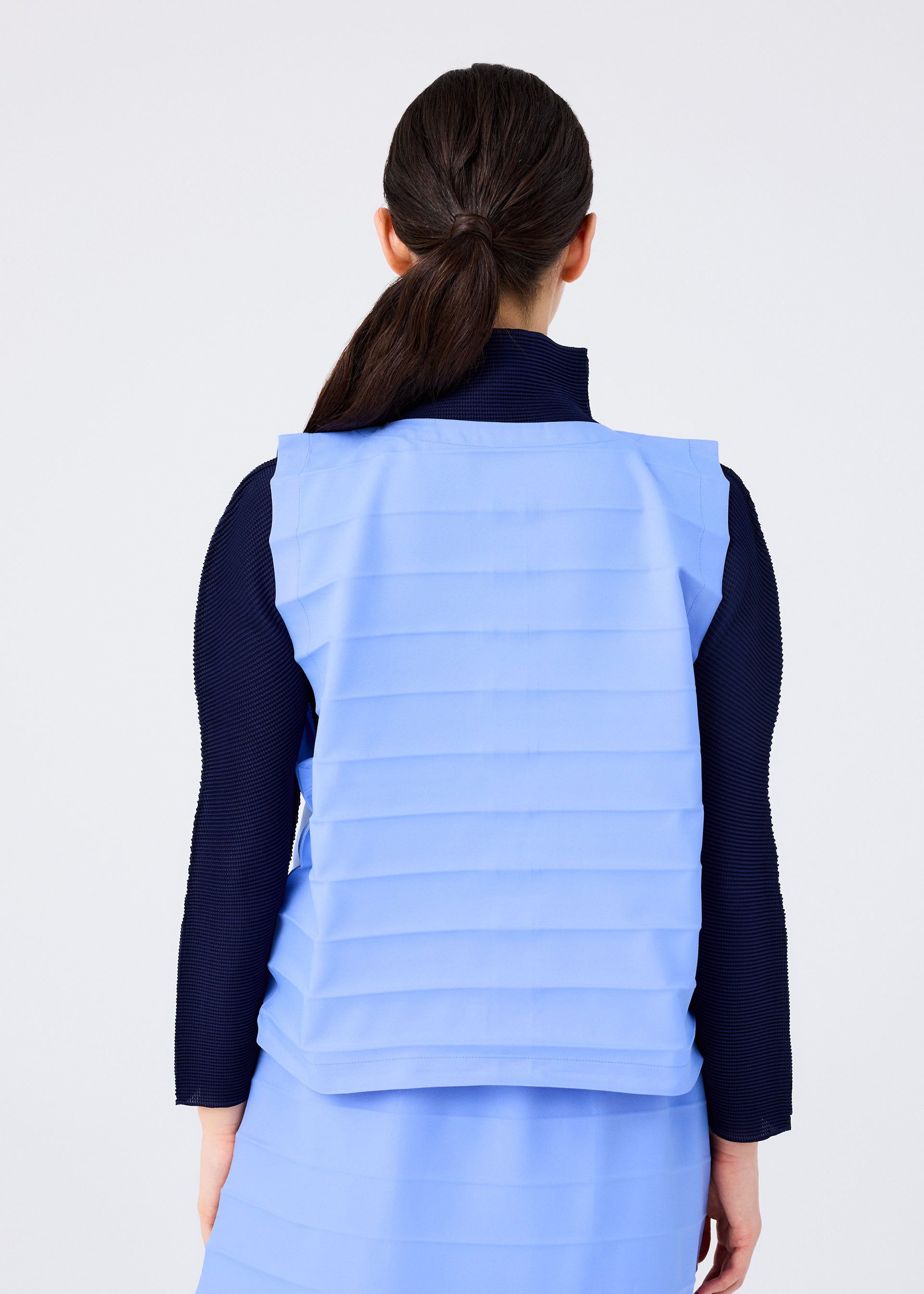Zoomed view A woman with dark hair in a low ponytail is seen from behind, wearing the light blue RIPPLE PLEATS top and matching skirt by me ISSEY MIYAKE over a long-sleeved, dark navy ribbed shirt against a plain white background.