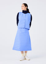 A woman stands against a white background wearing the light blue RIPPLE PLEATS vest and matching long skirt from me ISSEY MIYAKE over a black turtleneck, with her hands behind her back, white shoes, and light-colored socks.