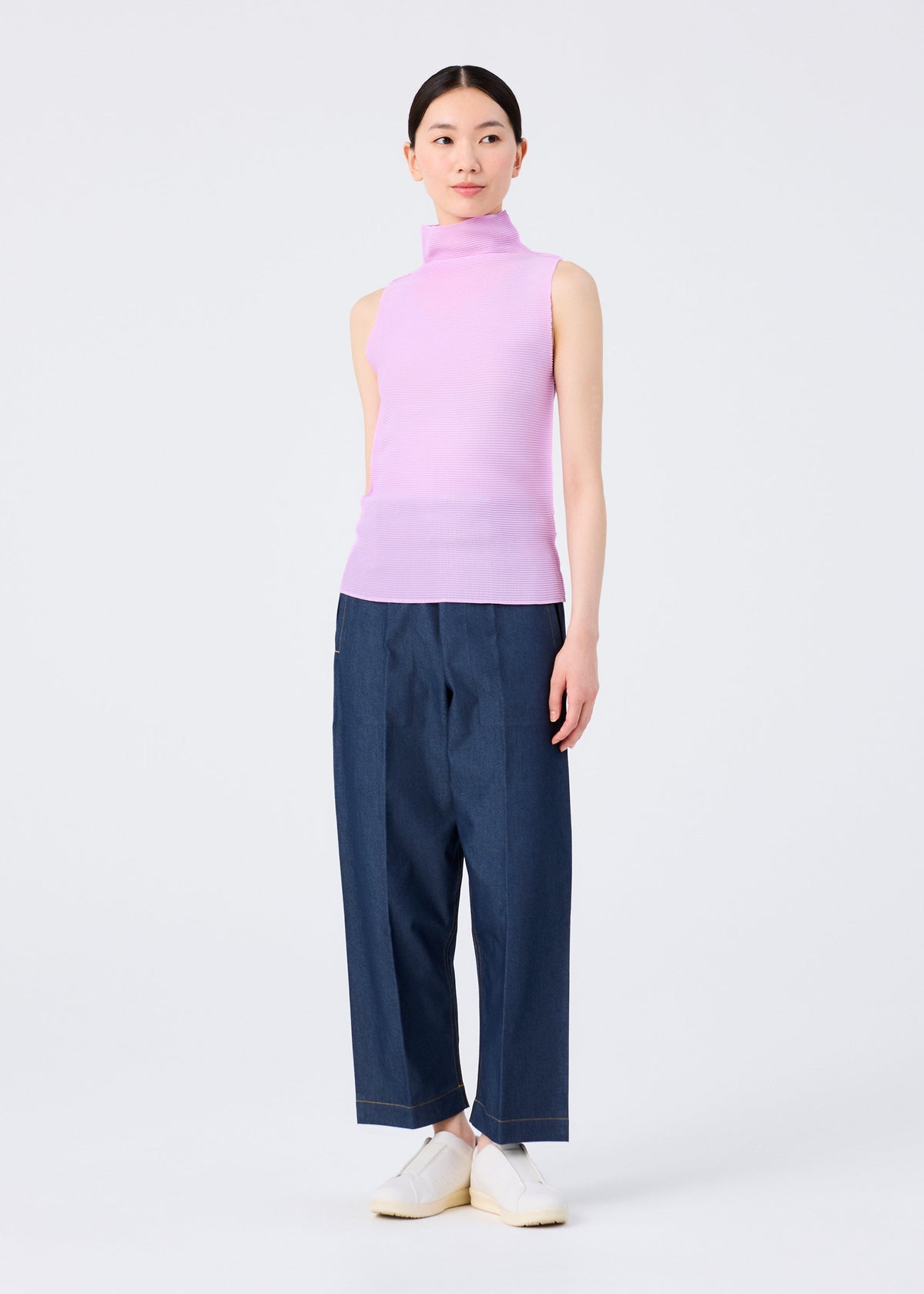 Against a plain white background, a person wears the me ISSEY MIYAKE STRETCH PLEATS 5 high-neck sleeveless light purple top, loose dark blue pants, and white slip-on shoes; their straight dark hair is pulled back. view 7 of 7