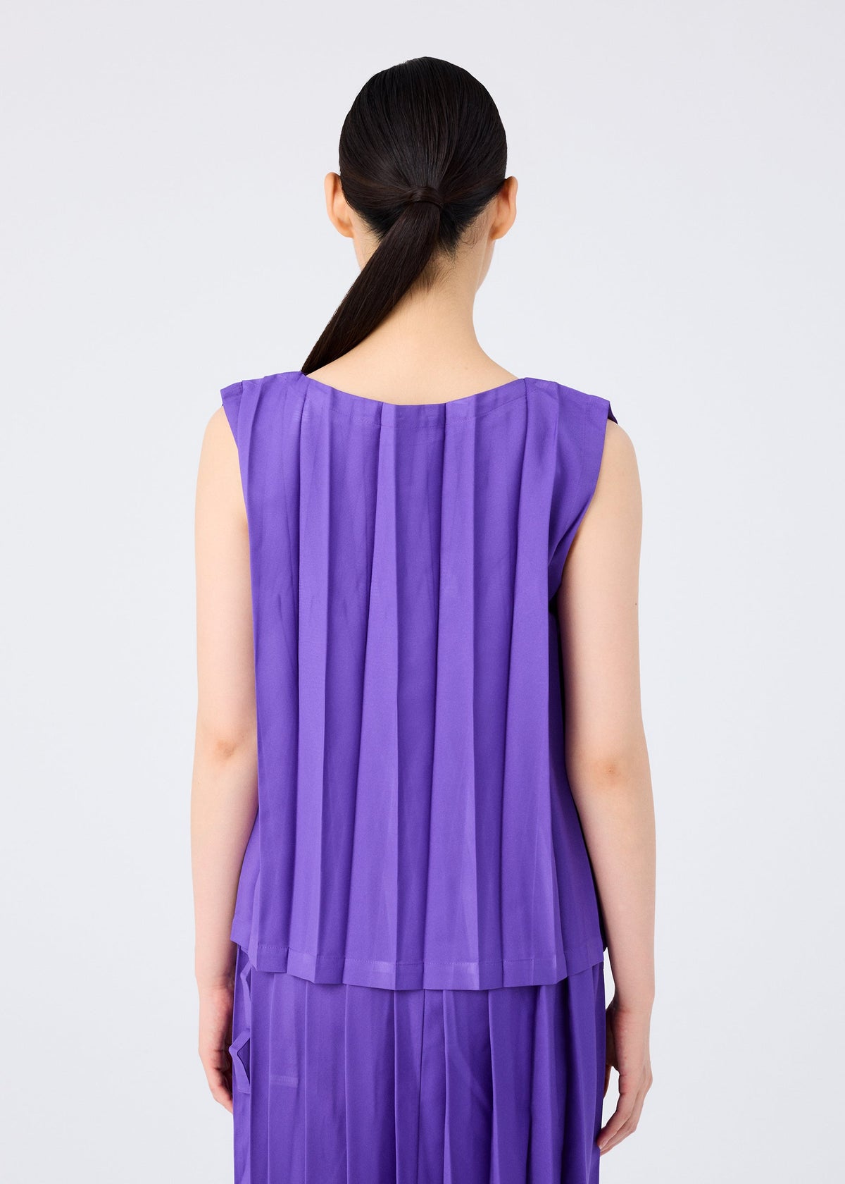 A person with long dark hair tied back faces away, wearing the me ISSEY MIYAKE CROSS PLEATS sleeveless top and skirt in rich purple stretch pleats. The plain, light-colored background highlights the textured design. view 7 of 8