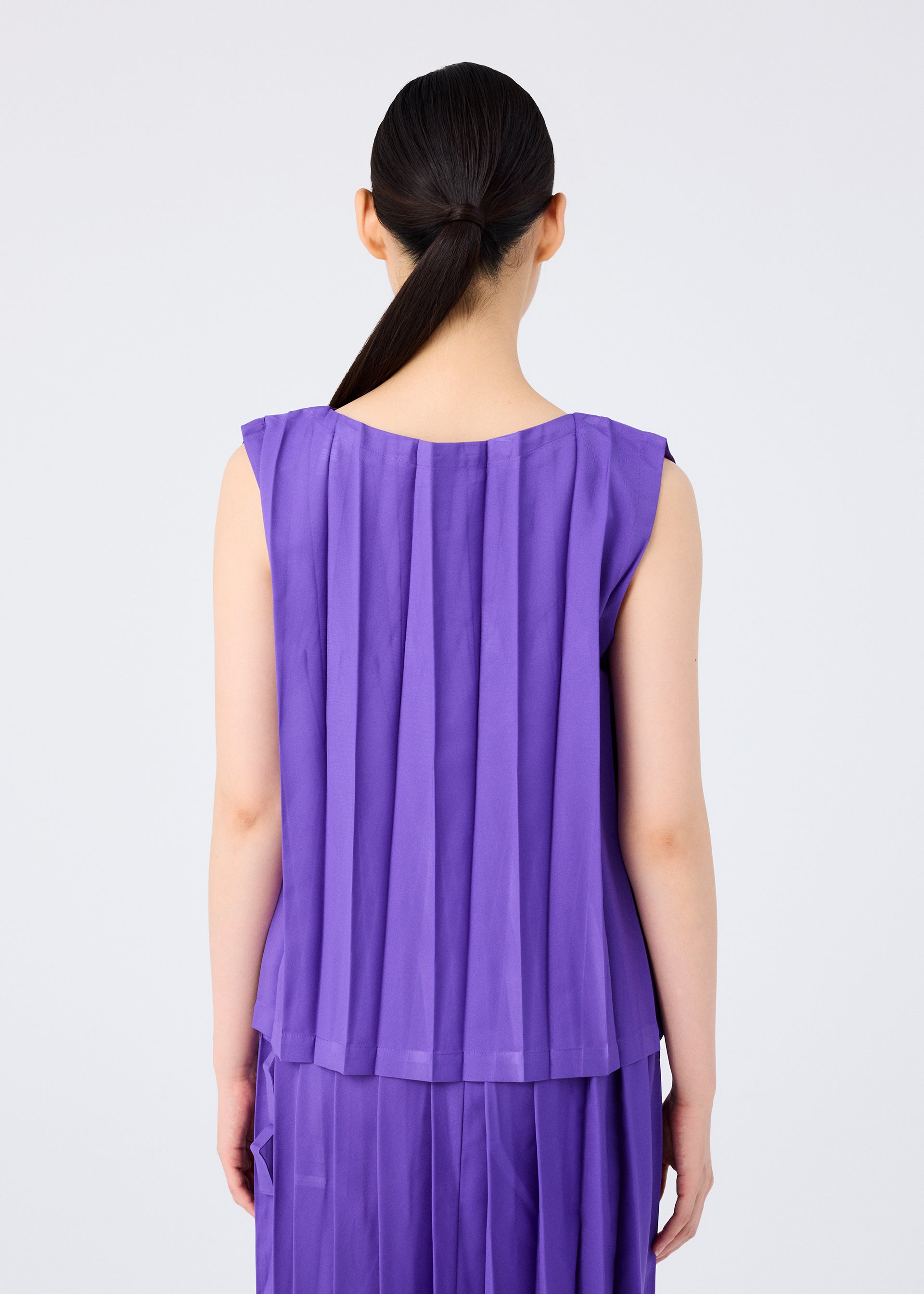 Zoomed view A person with long dark hair tied back faces away, wearing the me ISSEY MIYAKE CROSS PLEATS sleeveless top and skirt in rich purple stretch pleats. The plain, light-colored background highlights the textured design.