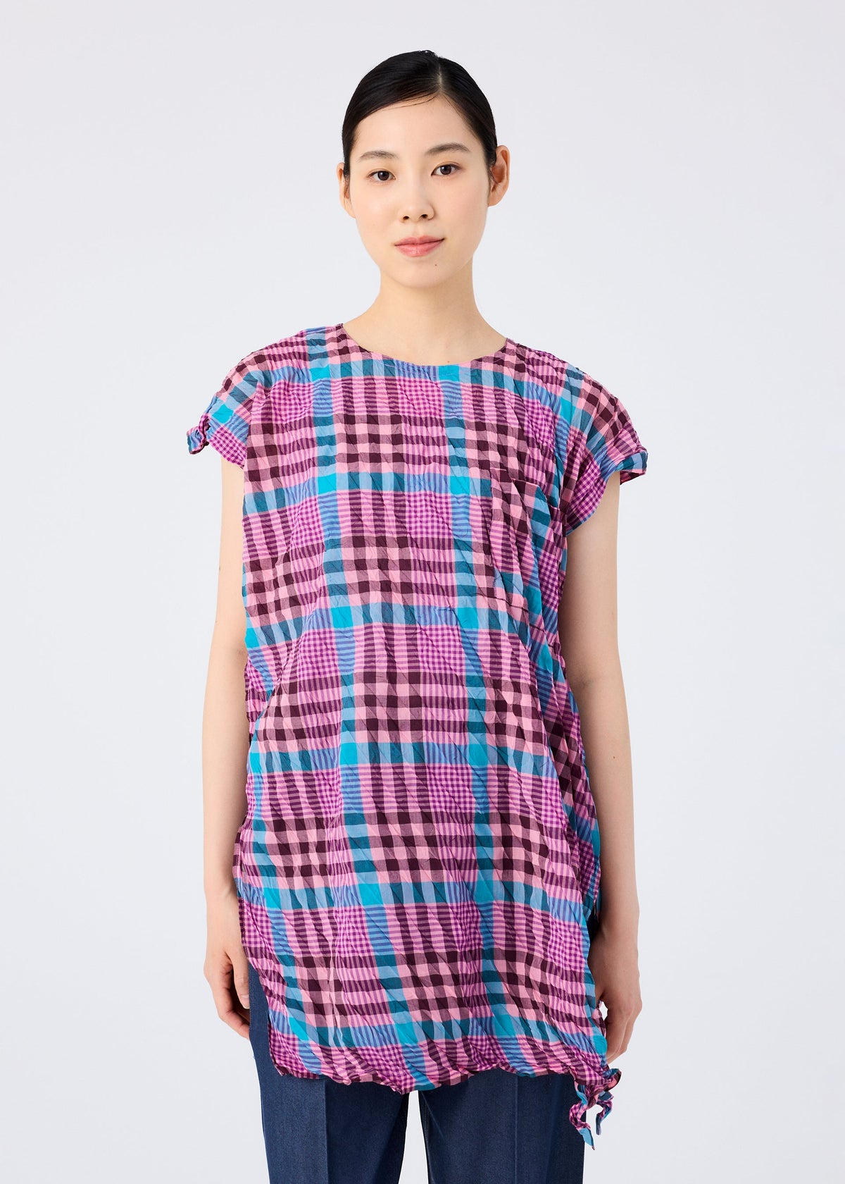 A woman with straight black hair, wearing the CHECK WHIPPED PLEATS SHIRT by me ISSEY MIYAKE in colorful pink, blue, and black checks with dark pants, stands facing forward against a plain light gray background with a neutral expression. view 5 of 7
