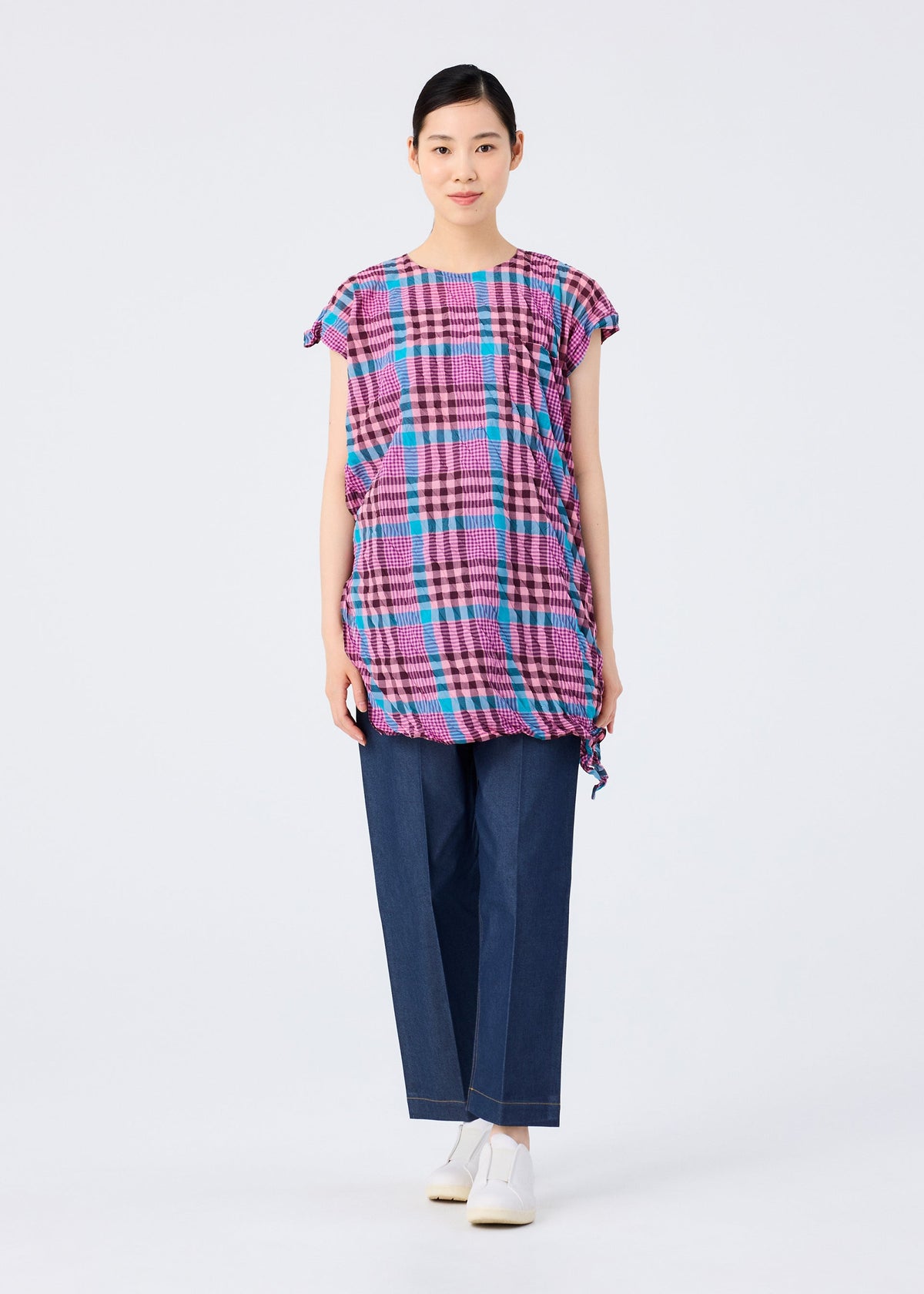 A person faces forward against a plain white background, wearing the me ISSEY MIYAKE CHECK WHIPPED PLEATS SHIRT in pink and blue checkered fabric with side ties, loose dark blue pants, and white shoes. view 7 of 7