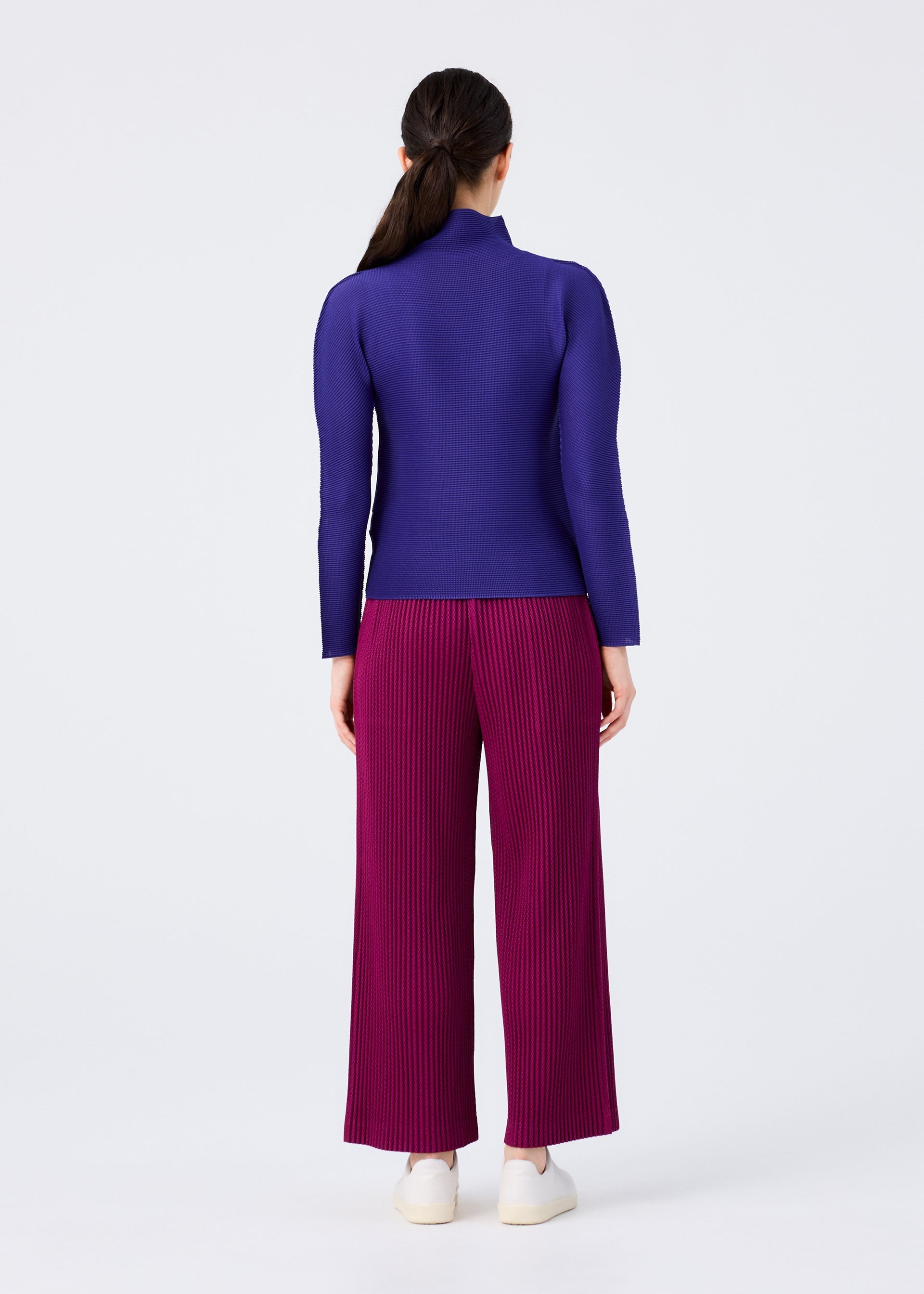Zoomed view Against a plain light background, a woman faces away wearing me ISSEY MIYAKE's WARM MESH PLEATS: a fitted purple top and wide-legged, pleated magenta pants with thick brushed mesh detailing, paired with white shoes.