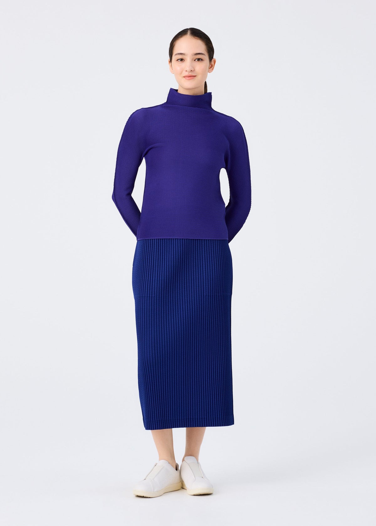 A woman stands facing forward against a white background, wearing the WARM MESH PLEATS blue midi skirt by me ISSEY MIYAKE, paired with a purple turtleneck and white slip-on shoes. Her hands are behind her back, and she is smiling slightly. view 5 of 7