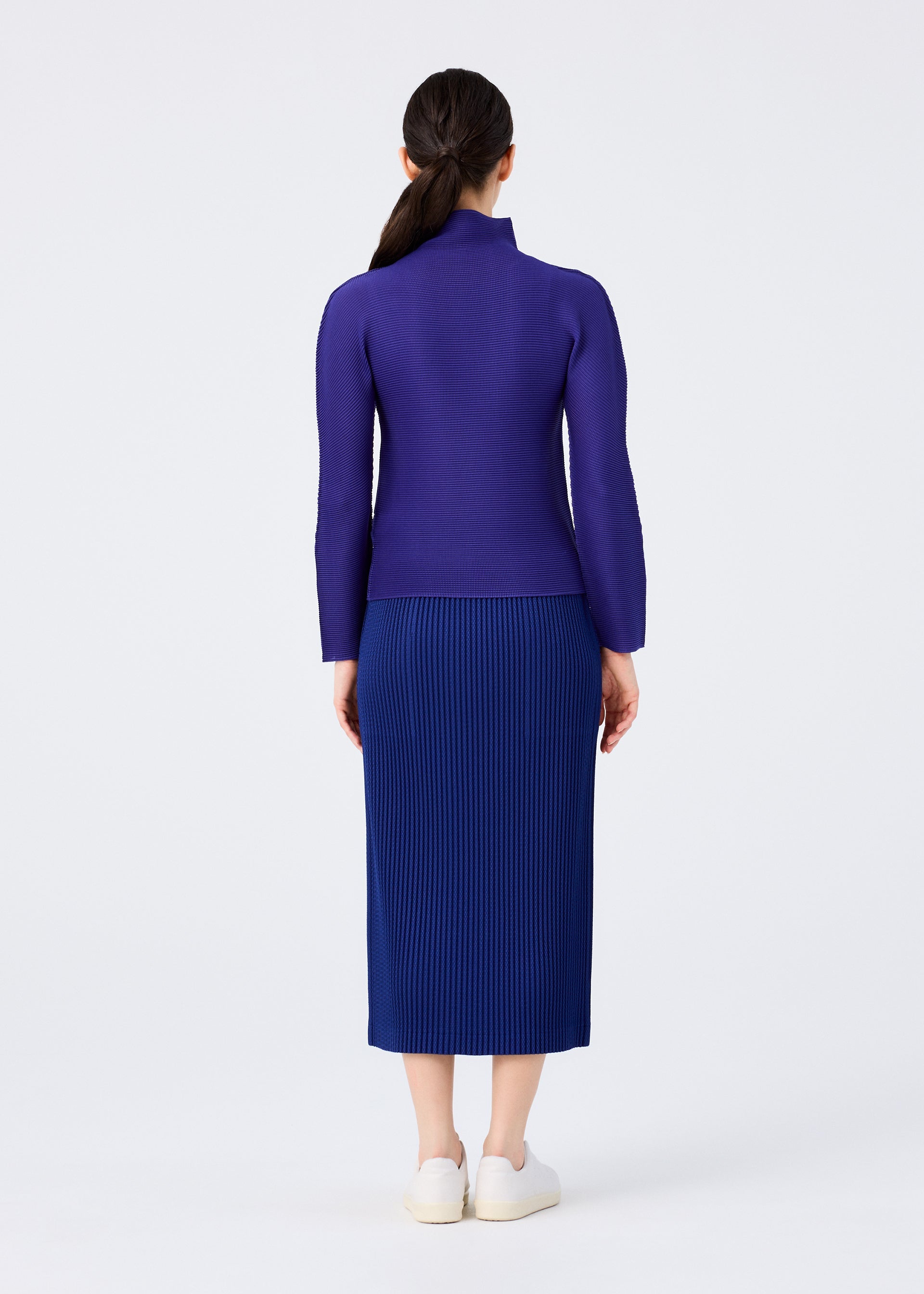 Zoomed view A woman faces away from the camera wearing the WARM MESH PLEATS blue midi skirt by me ISSEY MIYAKE, paired with a long-sleeved purple top and white slip-on shoes, against a plain white background.