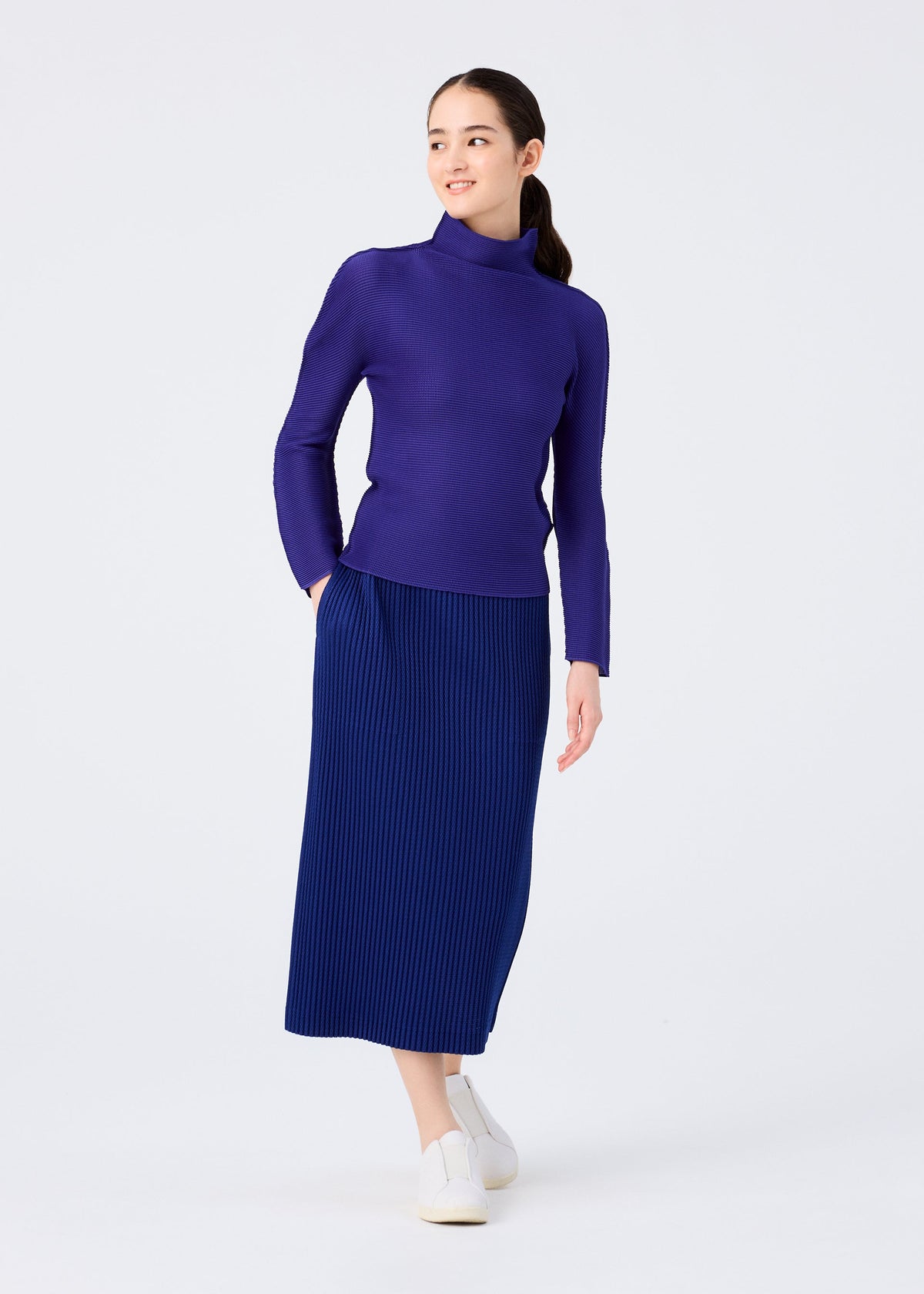 A woman stands against a white background, smiling to the side with one hand in her pocket, wearing the me ISSEY MIYAKE WARM MESH PLEATS skirt, a long-sleeve purple turtleneck sweater, and white slip-on shoes. view 7 of 7
