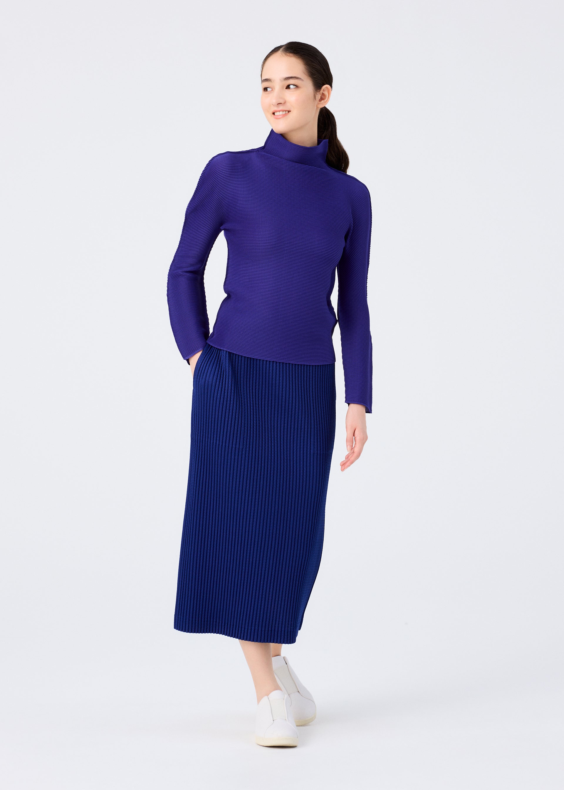 Zoomed view A woman stands against a white background, smiling to the side with one hand in her pocket, wearing the me ISSEY MIYAKE WARM MESH PLEATS skirt, a long-sleeve purple turtleneck sweater, and white slip-on shoes.