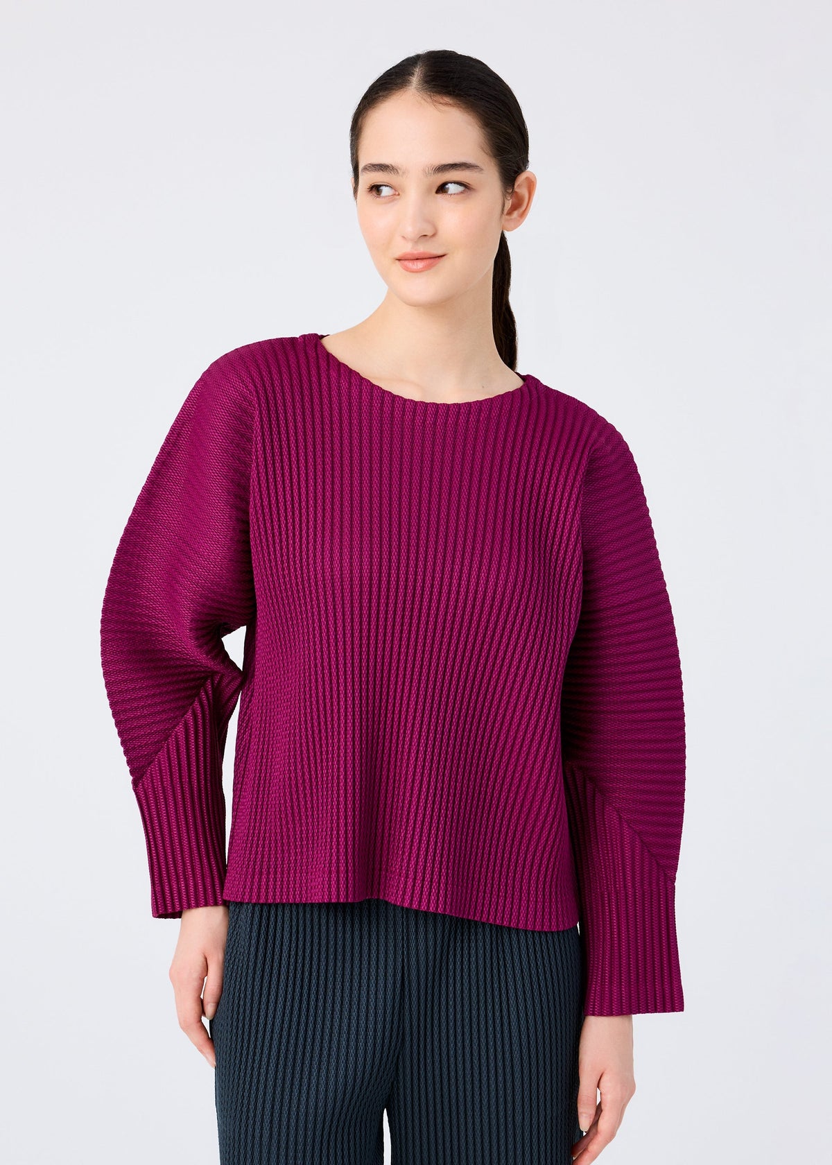 A woman with long dark hair wears the me ISSEY MIYAKE WARM MESH PLEATS top in textured magenta with pleats and dark ribbed pants, standing against a light gray background and smiling subtly. view 5 of 7
