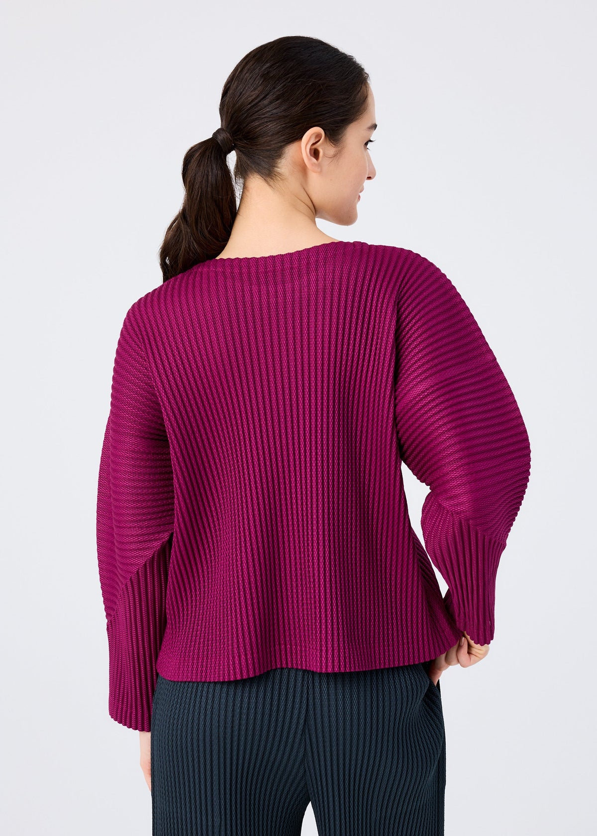 A woman with dark hair in a low ponytail is seen from the back, wearing the WARM MESH PLEATS top by me ISSEY MIYAKE and dark pinstriped pants against a plain light background. view 6 of 7