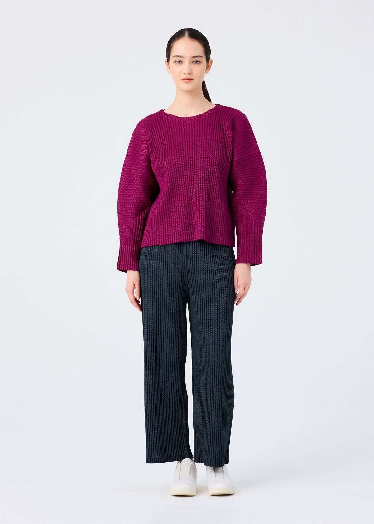 A person faces forward against a plain white background, dressed in the me ISSEY MIYAKE WARM MESH PLEATS—a loose, ribbed magenta long-sleeved sweater—paired with dark pinstriped wide-leg pants and white sneakers. view 7 of 7