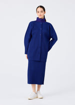 A woman poses against a white backdrop in the WARM MESH PLEATS set by me ISSEY MIYAKE, featuring a royal blue ribbed skirt and long-sleeved cardigan, with her hair pulled back and white slip-on shoes.