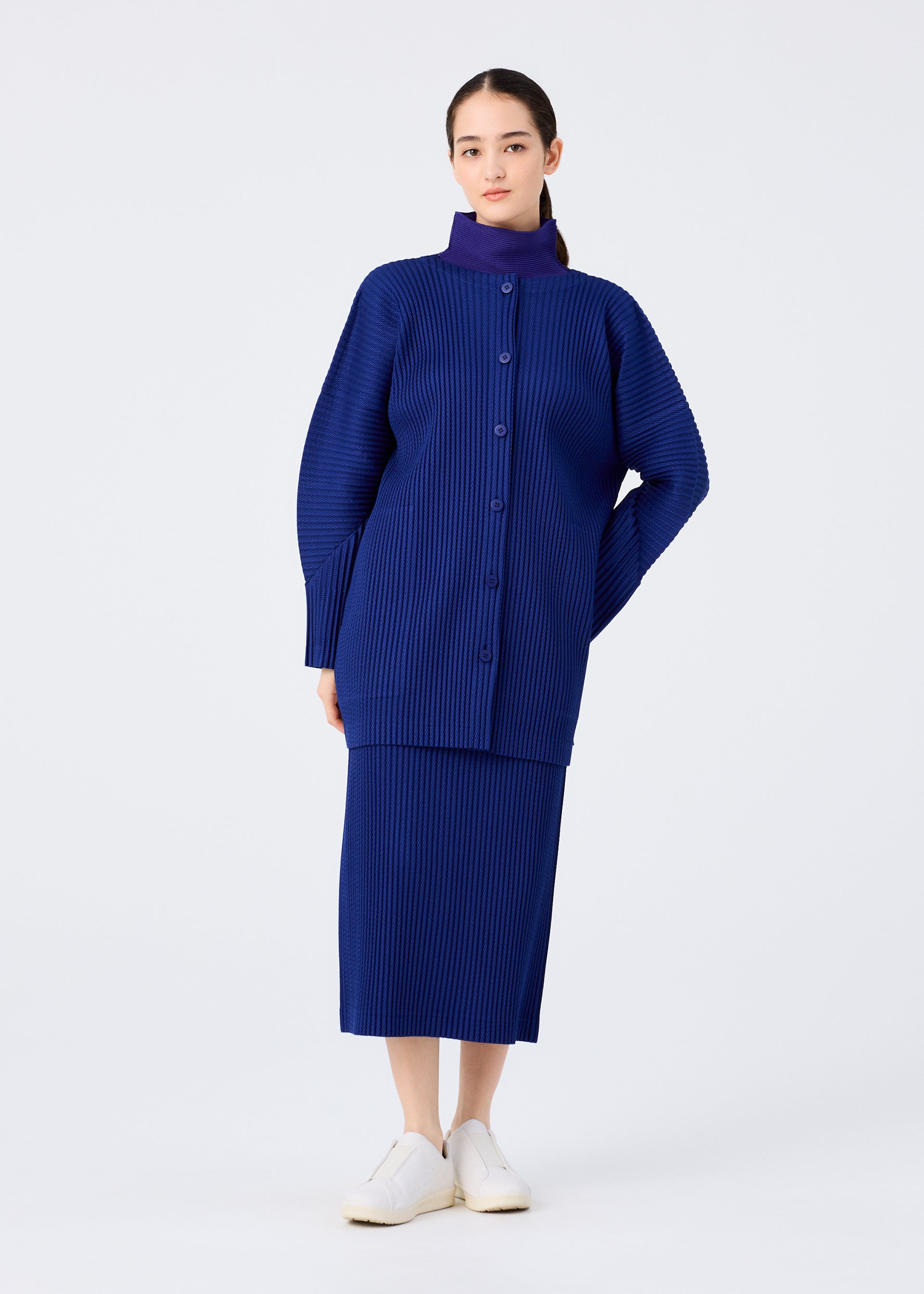 Zoomed view A woman poses against a white backdrop in the WARM MESH PLEATS set by me ISSEY MIYAKE, featuring a royal blue ribbed skirt and long-sleeved cardigan, with her hair pulled back and white slip-on shoes.