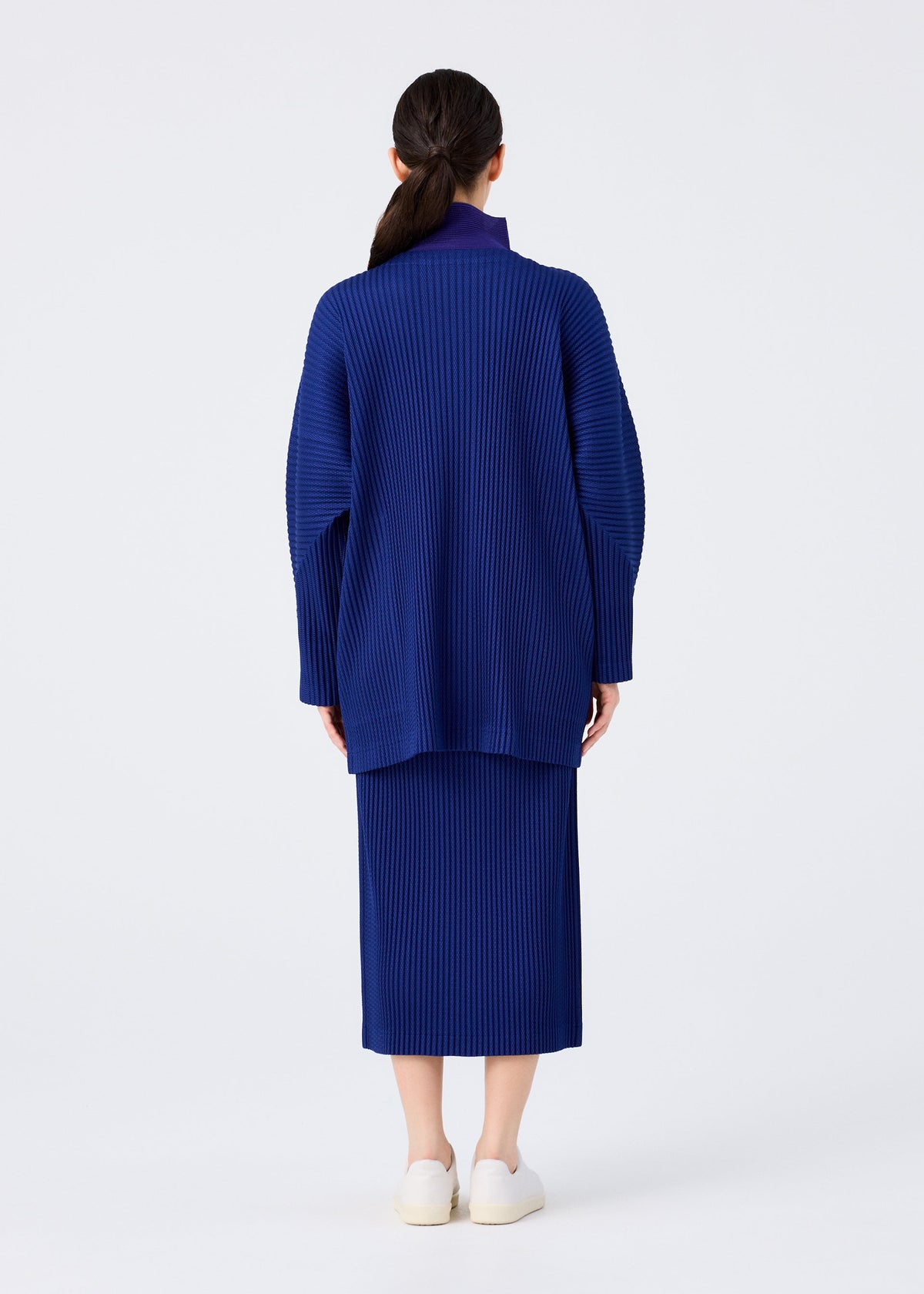 A woman faces away from the camera, wearing the WARM MESH PLEATS set by me ISSEY MIYAKE—a matching blue long-sleeved cardigan and skirt—with white slip-on shoes, posed against a plain white background. view 7 of 7
