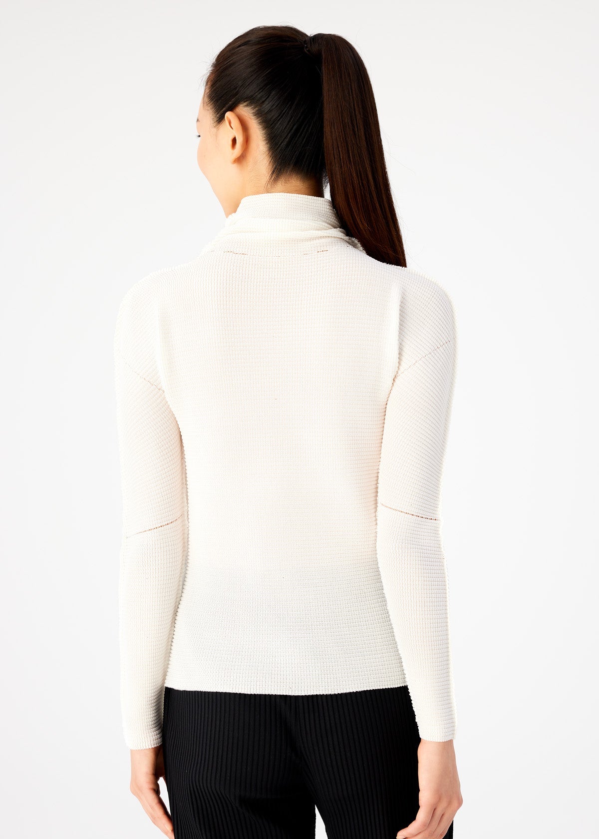 A woman with long dark hair in a ponytail is seen from behind, wearing a white KNIT STRETCH PLEATS top from me ISSEY MIYAKE and black pants against a plain light background. view 5 of 6