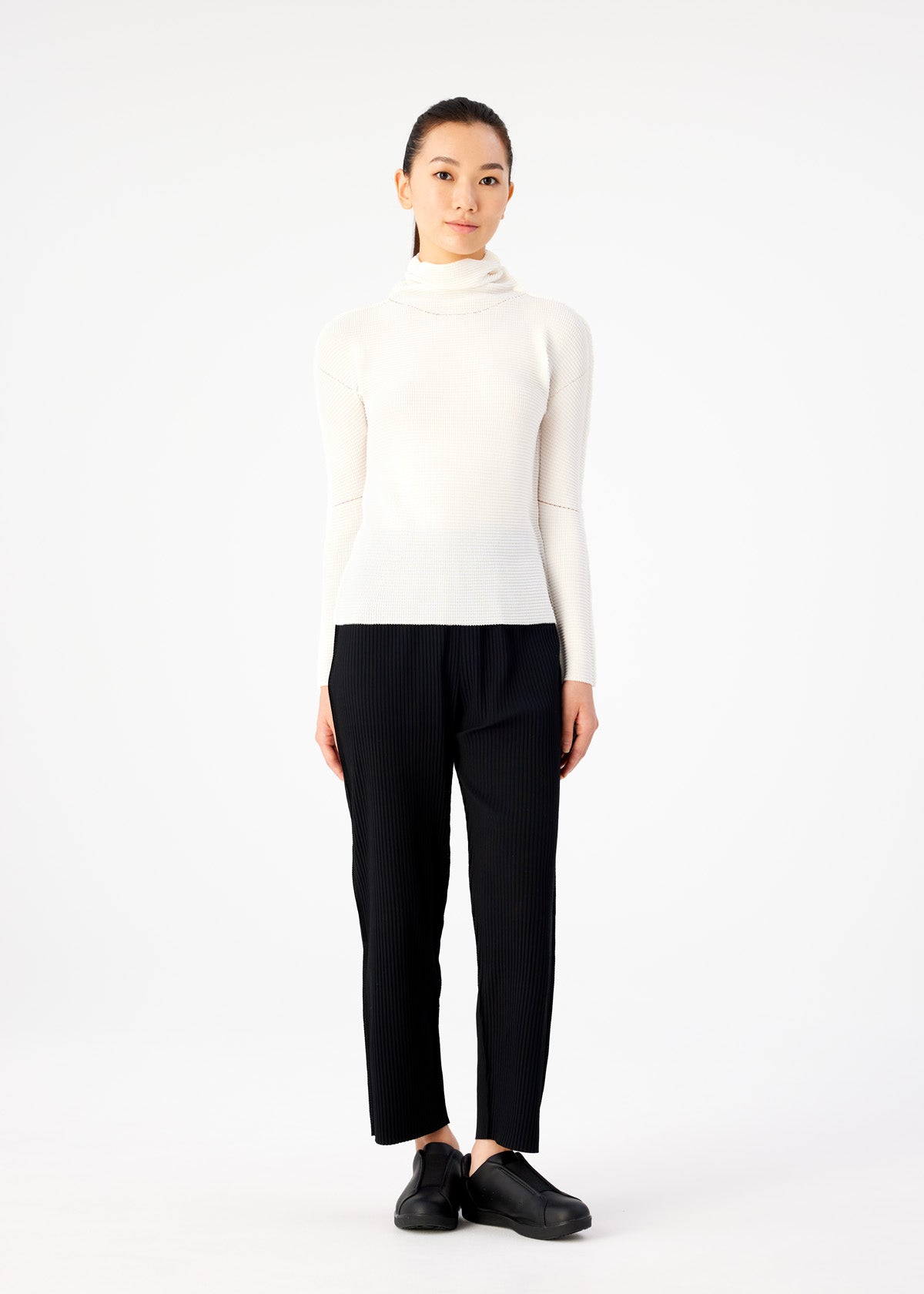 Zoomed view A woman stands facing forward against a plain white background, wearing the KNIT STRETCH PLEATS top by me ISSEY MIYAKE with black pants and shoes. Her hands are relaxed at her sides and she has a neutral expression.