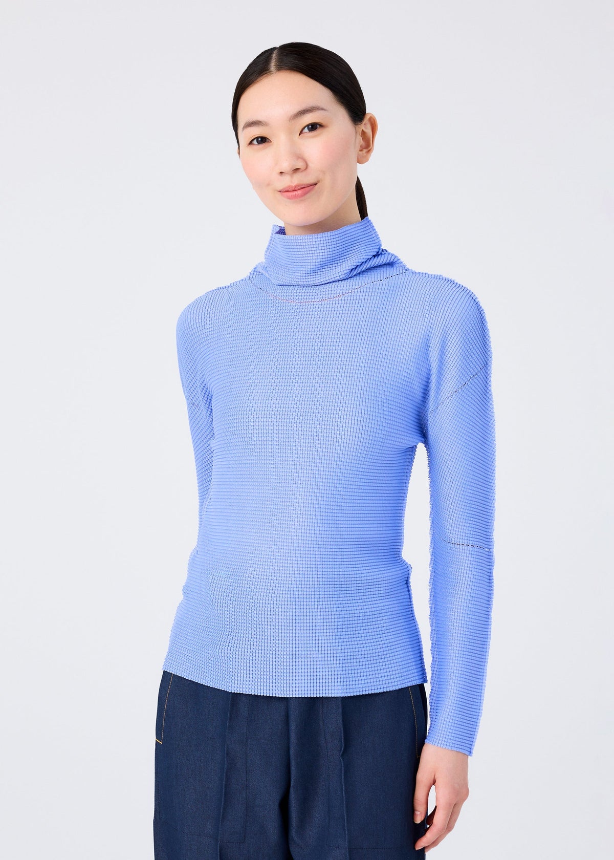 A woman with straight dark hair pulled back wears the me ISSEY MIYAKE KNIT STRETCH PLEATS 1 top in light blue with long sleeves and a high neck, paired with dark pants, standing against a white background and smiling gently. view 5 of 7