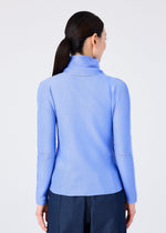 A woman with long dark hair in a low ponytail faces away, wearing the me ISSEY MIYAKE KNIT STRETCH PLEATS 1 top in light blue with long sleeves and a high neck, paired with dark pants against a plain light background.