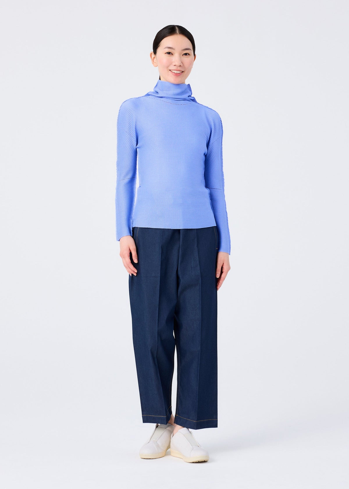 A person stands smiling against a plain white background, wearing the KNIT STRETCH PLEATS 1 by me ISSEY MIYAKE—a light blue long sleeve high neck pleated top—styled with wide-leg dark blue pants and white slip-on shoes. view 7 of 7