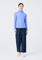 A person stands smiling against a plain white background, wearing the KNIT STRETCH PLEATS 1 by me ISSEY MIYAKE—a light blue long sleeve high neck pleated top—styled with wide-leg dark blue pants and white slip-on shoes.