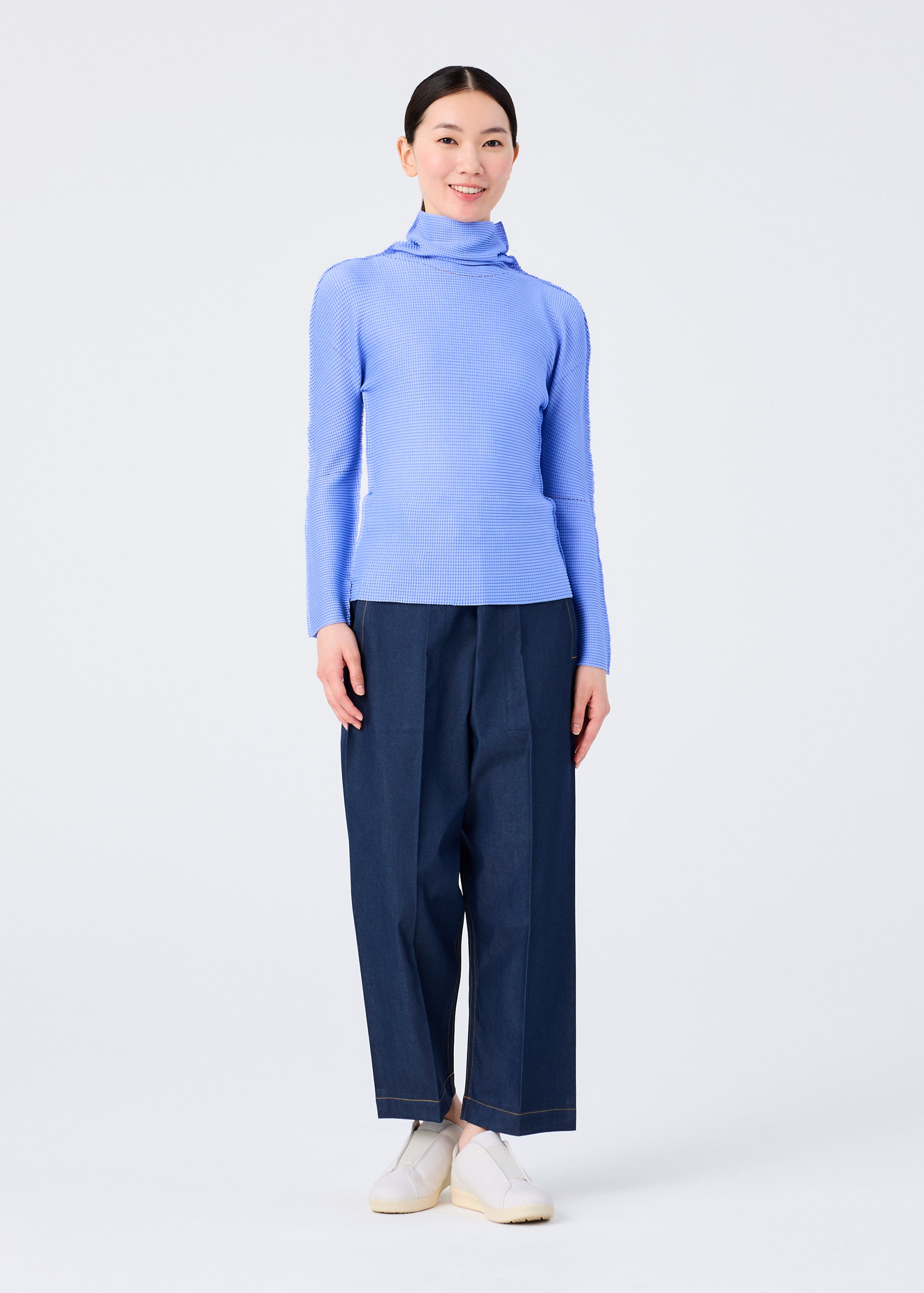 Zoomed view A person stands smiling against a plain white background, wearing the KNIT STRETCH PLEATS 1 by me ISSEY MIYAKE—a light blue long sleeve high neck pleated top—styled with wide-leg dark blue pants and white slip-on shoes.