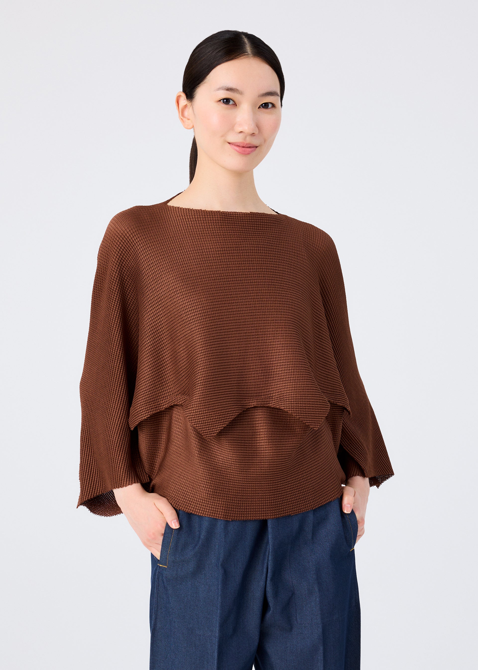 Zoomed view A woman with dark hair tied back is wearing the me ISSEY MIYAKE GIZA GIZA KNIT STRETCH PLEATS top in brown, paired with blue pants, standing against a plain light background and smiling softly at the camera.