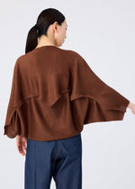 A woman faces away wearing the me ISSEY MIYAKE GIZA GIZA KNIT STRETCH PLEATS top in brown with wide sleeves and blue pants. Her dark hair is tied back in a low ponytail as she turns slightly to the right.