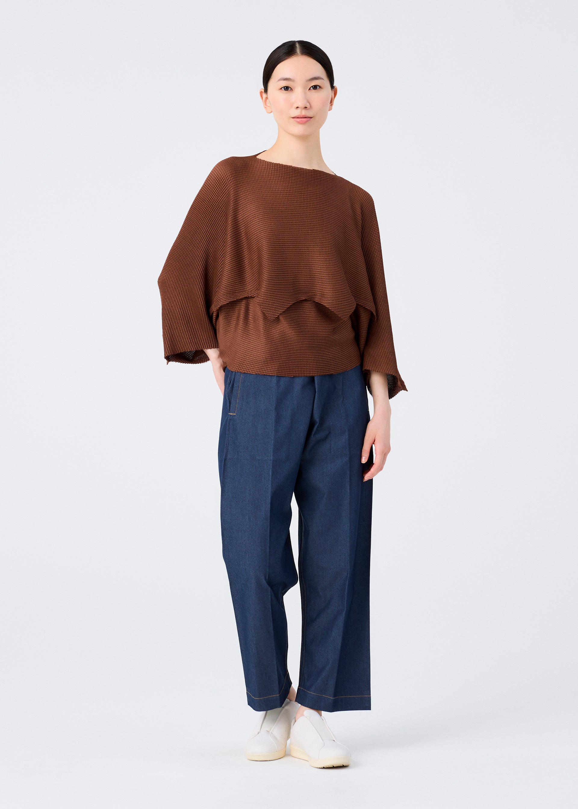 Zoomed view A woman with straight hair pulled back and a neutral expression models me ISSEY MIYAKE's GIZA GIZA KNIT STRETCH PLEATS: a loose brown asymmetric 3/4-sleeve top, blue wide-leg pants with visible stitching, and white slip-on shoes against a white background.
