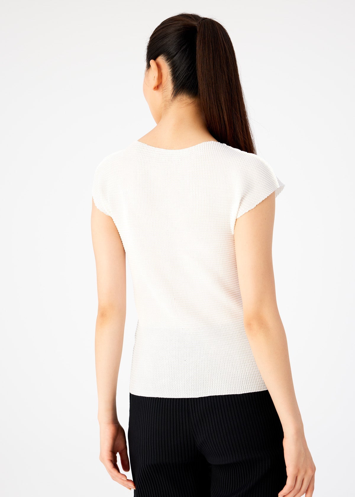 Zoomed view A woman with long dark hair in a ponytail is shown from the back, wearing me ISSEY MIYAKE’s KNIT STRETCH PLEATS round neck white top with French sleeves and black pants against a plain light background.
