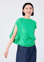 A person with short black hair smiles, wearing the me ISSEY MIYAKE GIZA GIZA KNIT STRETCH PLEATS—a bright green, French-sleeve knit top with side slits—paired with blue wide-leg pants against a light background.