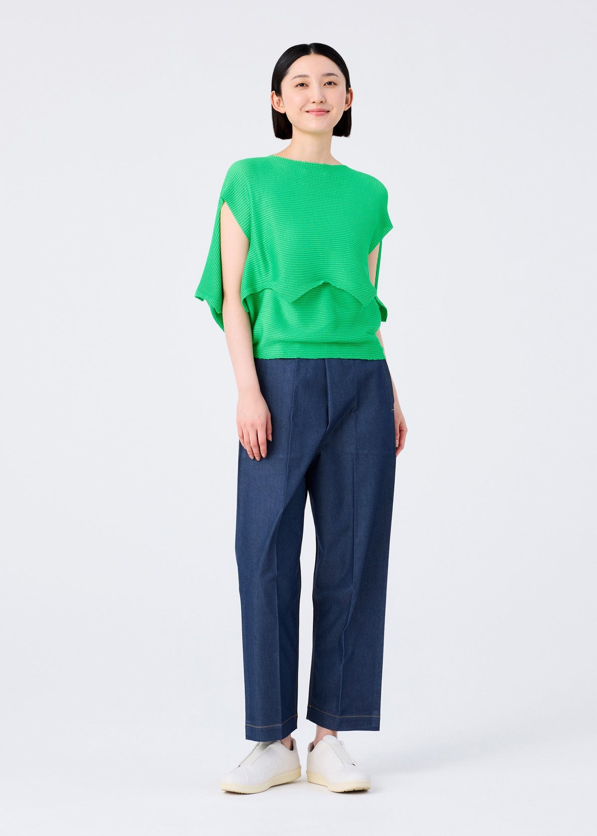 A person stands facing forward in a bright green me ISSEY MIYAKE GIZA GIZA KNIT STRETCH PLEATS top, dark blue ankle-length pants, and white slip-on shoes against a plain white background. view 8 of 9