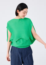 A person with short dark hair wears the me ISSEY MIYAKE GIZA GIZA KNIT STRETCH PLEATS bright green poncho-style top and blue wide-leg pants, standing against a plain light background, looking down and smiling.